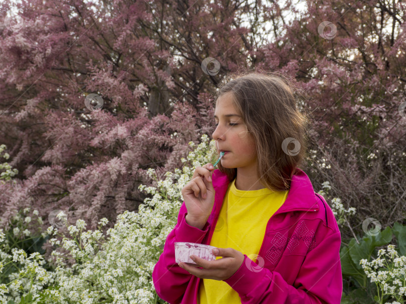 Скачать Девочка-подросток наслаждается вкусом десерта. Девочка ест мороженое из пластиковой баночки и счастливо закрывает глаза на прогулке на фоне цветущих деревьев и растений фотосток Ozero