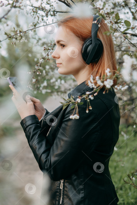 Скачать Рыжеволосая девочка-подросток слушает музыку в наушниках, настроенных с помощью телефона. Время весеннего цветения вишневых садов фотосток Ozero
