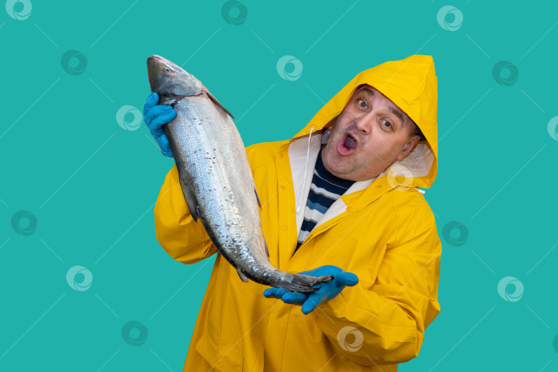 Скачать рыболов в желтом костюме держит в руках свежевыловленную рыбу. рыбак удивлен хорошим уловом. рыбак на фоне моря. ловец в плаще. фотосток Ozero