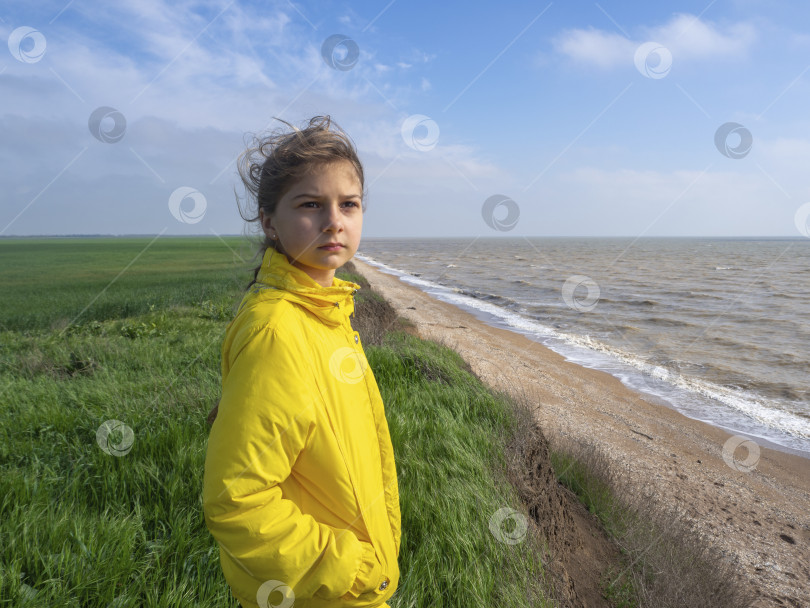 Скачать Девушка в желтой ветровке смотрит вдаль, стоя на зеленом травянистом лугу у высокого берега на фоне голубого неба и серого моря фотосток Ozero