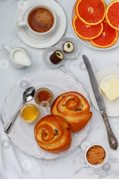 Скачать Две булочки с изюмом, джемом и медом на белой тарелке на мраморном фоне на завтрак. Кофе, молоко, сливочное масло, арахисовое масло, шоколад и апельсины на столе. Выборочный фокус, вид сверху фотосток Ozero