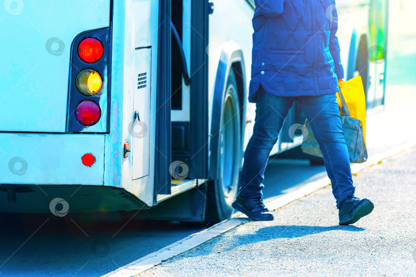 Скачать Белый городской автобус с открытыми дверями на автовокзале, из которого выходит не в фокусе внимания пассажир с сумками в руках. Снимок сделан с подсветкой и солнечными бликами. фотосток Ozero