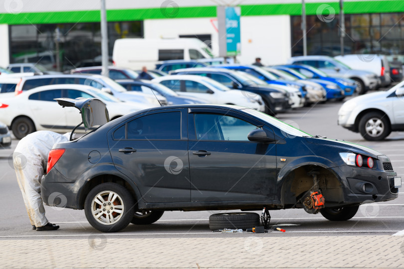 Скачать Автомобиль со снятым колесом стоит на домкрате на стоянке супермаркета. Мужчина-водитель достает из багажника инструмент, чтобы починить спущенную шину. фотосток Ozero