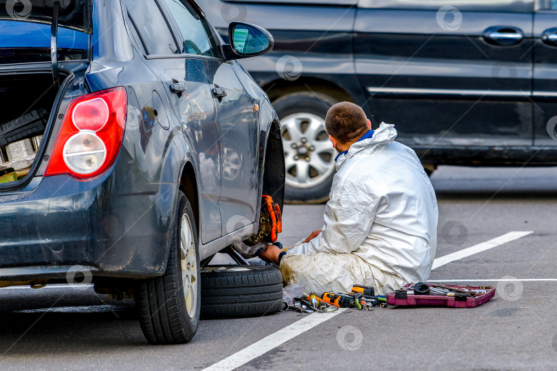 Скачать Автомобиль со снятым колесом стоит на домкрате. Мужчина-водитель чинит спущенную шину на своем автомобиле. фотосток Ozero