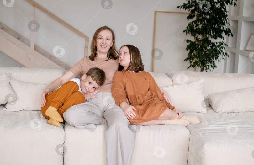 Скачать Портрет матери с детьми. Женщина, сидящая на диване с сыном и дочерью на фоне домашнего интерьера. Семья отдыхает вместе на удобном диване в гостиной. фотосток Ozero