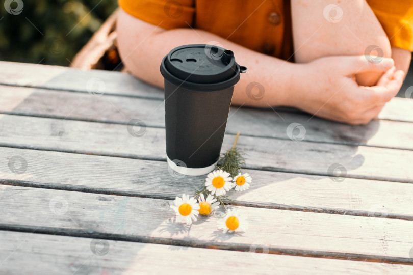 Скачать Черный бумажный стаканчик для кофе, стоящий на деревянном столе с веточкой ромашек и женскими руками на заднем плане фотосток Ozero