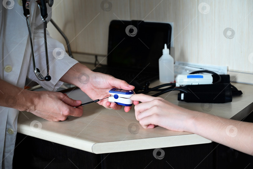 Скачать Кислородный тест на covid. Врач осматривает пациента с помощью пульсоксиметра на кончике пальца за деревянным столом, крупным планом. фотосток Ozero