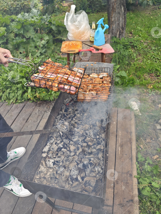 Скачать Приготовление шашлыка на мангале на природе. молодой человек переворачивает угли и ставит решетку с барбекю на решетку гриля. на заднем плане видны трава и деревья фотосток Ozero