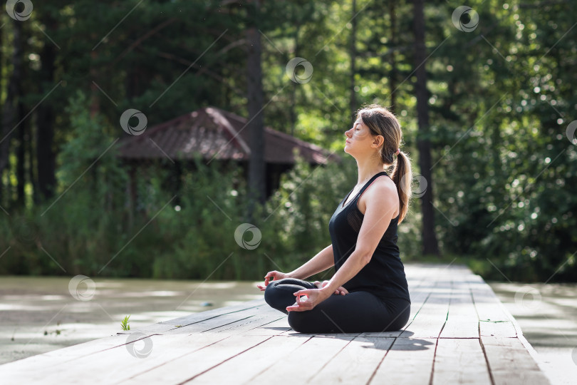 Скачать Женщина практикует медитацию, сидя в позе лотоса на деревянном мосту в парке летним солнечным утром, занимается йогой в черной спортивной одежде фотосток Ozero
