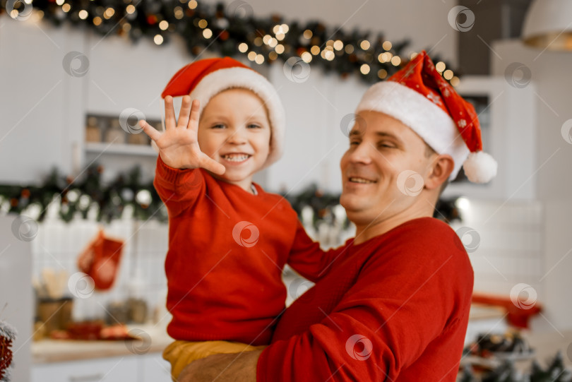 Скачать Концепция "Счастливого Рождества" и "Счастливого Нового года". Счастливый отец держит на руках своего милого улыбающегося сына, смотрящего в камеру и показывающего пять пальцев на ладони на празднично украшенной кухне. Оба мужчины одеты в красные свитера и шапочки Санты фотосток Ozero
