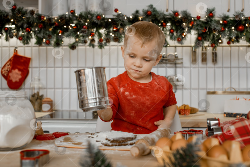 Скачать Милый маленький мальчик очень занят просеиванием муки через сито для теста на рождественски украшенной кухне у себя дома фотосток Ozero