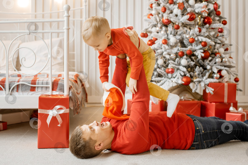 Скачать Улыбающийся малыш в красном свитере, летящий на руках у своего отца со счастливым смешным лицом на фоне новогодней елки и рождественских подарков фотосток Ozero