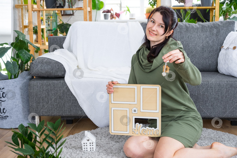 Скачать Счастливая женщина в доме держит в руках фоторамку с рисунком по шаблону дома и ключом в интерьере. Проект дома мечты, покупка недвижимости, страхование, ипотека, аренда, бронирование фотосток Ozero