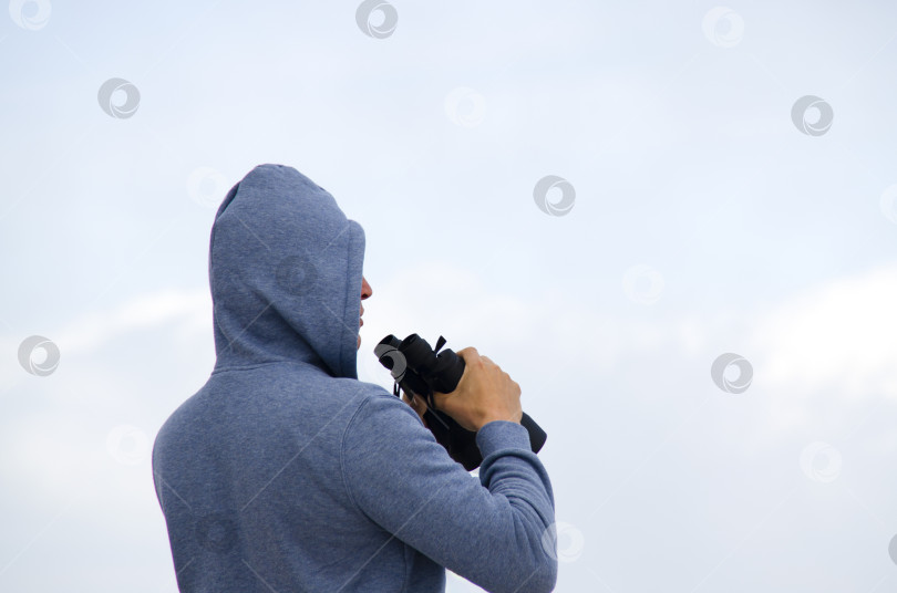 Скачать мужчина в синей куртке с капюшоном на голове смотрит вдаль в бинокль. Мужчина на фоне голубого неба. фотосток Ozero