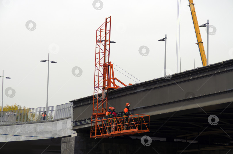 Скачать Рабочие в оранжевой униформе поднимаются на строительном лифте на вершину моста. Реконструкция, реставрация и ремонт объекта. фотосток Ozero