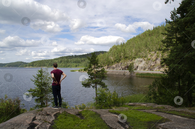 Скачать Молодой человек стоит, любуясь прекрасным видом. Озеро, ели на холмах, голубое небо с облаками. Северные пейзажи в Карелии, Россия. фотосток Ozero