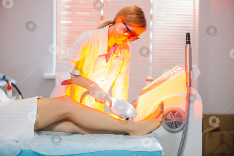 Скачать Свет, лазерная вспышка. Фотография симпатичной женщины-врача в специальных защитных очках, которая проводит процедуру лазерной эпиляции, удаление волос на ногах современными методами фотосток Ozero