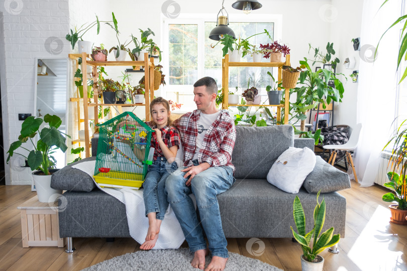 Скачать Неполная семья: папа, дочь и домашний попугай в клетке наслаждаются новым домом, сидя на диване. Ипотека, страхование и защита, покупка и переезд в собственный дом, зеленый домик с растением в горшке фотосток Ozero