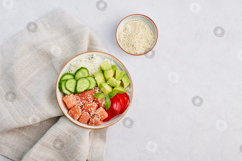 Скачать Выложите миску с лососем, рисом, авокадо, огурцом, перцем и кунжутом на льняную салфетку на сером фоне. фотосток Ozero