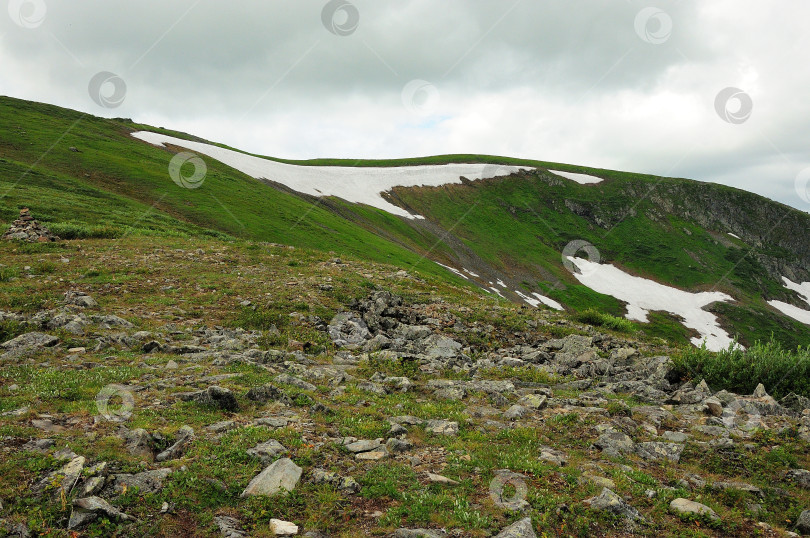 Скачать Вершина скалистого холма с остатками снега на склонах под пасмурным летним небом. фотосток Ozero