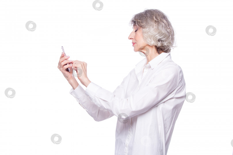 Скачать Красивая зрелая седовласая женщина улыбается с помощью смартфона, просматривая видео или общаясь в чате, отправляя текстовые сообщения, изолированные на белом фоне фотосток Ozero