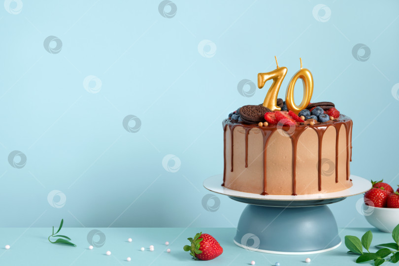 Скачать Шоколадный праздничный торт с ягодами, печеньем и числом семьдесят золотых свечей на синем фоне стены, копировальное пространство фотосток Ozero