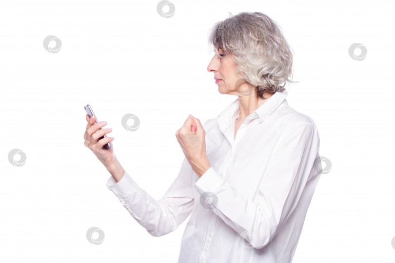 Скачать Приятная пожилая женщина выглядит очень сердитой и раздраженной, разговаривая по мобильному телефону. Потрясает кулаком телефонным мошенникам, изолированным на белом фоне фотосток Ozero