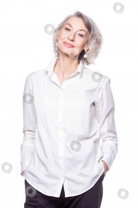 Скачать Счастливая модная пожилая женщина с красивой улыбкой, одетая в модную рубашку, стоит, засунув руки в карманы, и смотрит в камеру, изолированную на белом фоне фотосток Ozero