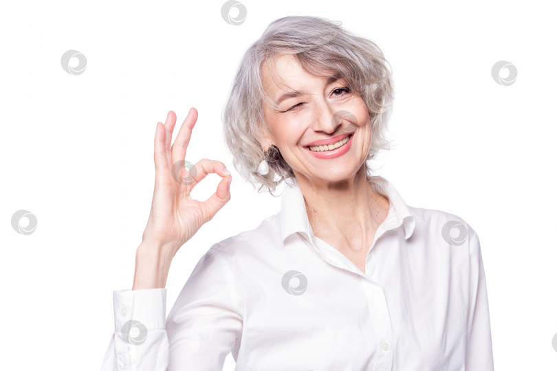 Скачать Крупным планом портрет зрелой привлекательной женщины в элегантной рубашке, стоящей на изолированном белом фоне, улыбающейся со счастливым лицом, подмигивающей в камеру, делающей знак "хорошо" фотосток Ozero
