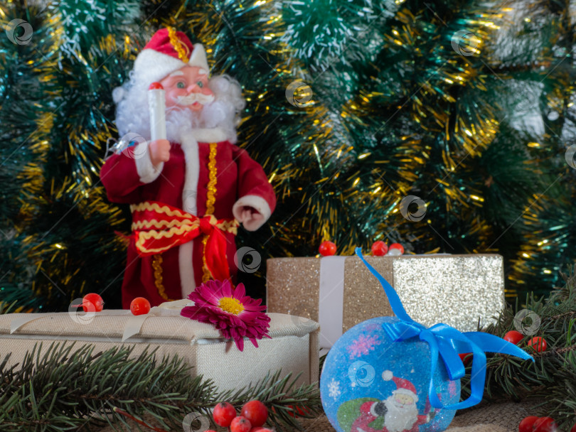 Скачать Рождественские подарки и украшения на фоне разноцветной зеленой мишуры. Золотая подарочная коробка из фактурной ткани с цветком, рождественским шаром, еловыми ветками, ягодами фотосток Ozero