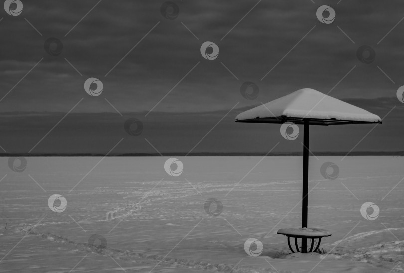 Скачать Одинокая скамейка с навесом на заснеженном пляже Северного моря.Зимний пейзаж в черно-белых тонах фотосток Ozero