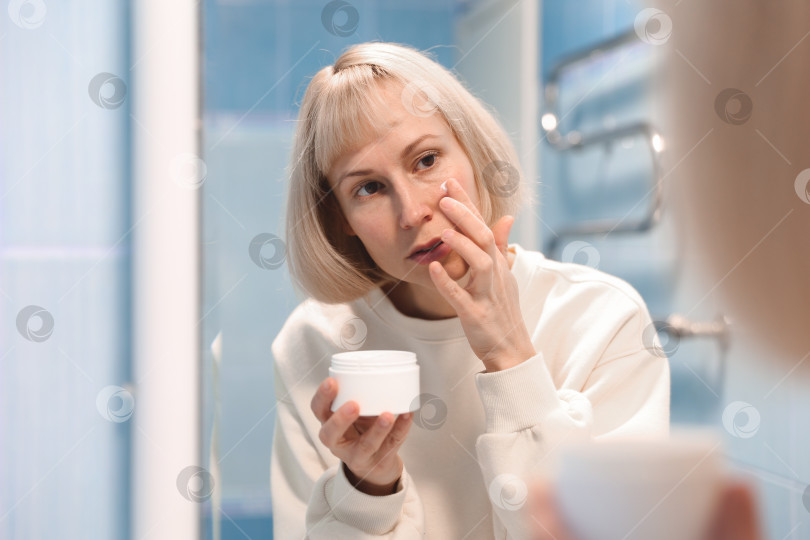 Скачать Красивая молодая женщина наносит увлажняющий крем на лицо в ванной, заботясь о своей коже фотосток Ozero