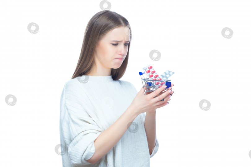 Скачать Студийный снимок расстроенной разочарованной девушки, держащей в руках маленькую тележку для покупок, полную дорогих таблеток и лекарств, выделенных на белом фоне фотосток Ozero