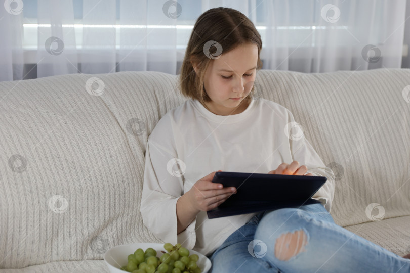 Скачать Умная маленькая девочка пользуется планшетным компьютером, сидя на диване в гостиной дома фотосток Ozero