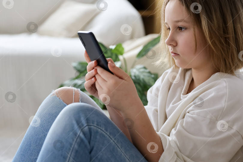 Скачать Красивая маленькая девочка играет в игру или смотрит видео на мобильном смартфоне. Девушка смотрит мультфильмы или просматривает Интернет. Портрет маленькой девочки, использующей смартфон сидя. фотосток Ozero