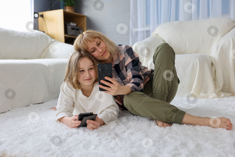 Скачать Стильная модная мать и дочь со смартфоном в руках снимают автопортрет на фронтальную камеру в гостиной, наслаждаясь свободным временем фотосток Ozero