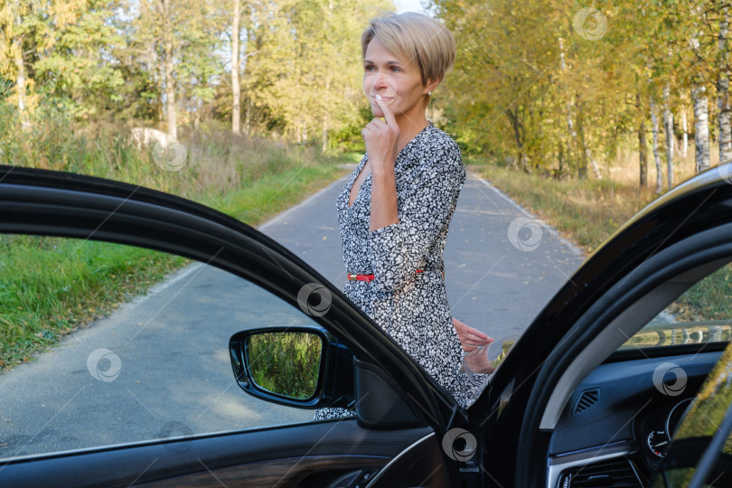 Скачать женщина в платье стоит перед автомобилем с открытой дверцей на фоне дороги и осенних деревьев фотосток Ozero