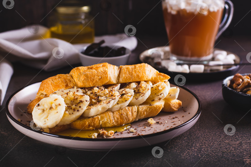 Скачать Вкусный круассан с бананом, медом, шоколадом и орехами на тарелке на столе фотосток Ozero