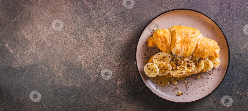 Скачать Бутерброд с круассаном с бананом, медом и орехами на тарелке на столешнице посмотреть веб-баннер фотосток Ozero