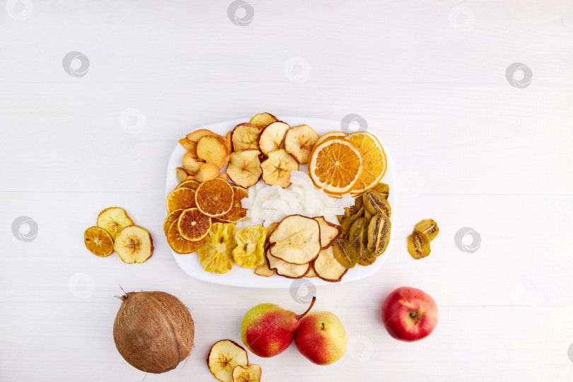 Скачать Сушеные яблоки, груши, ананасы, киви, абрикосы, кокосы, апельсины и мандариновые чипсы на тарелке на белом деревянном фоне. фотосток Ozero