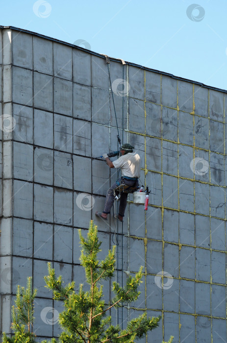 Скачать Промышленный альпинист ремонтирует стену высотного здания. Рабочий зачищает швы перфоратором и заполняет их строительной пеной. фотосток Ozero