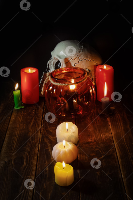 Скачать Натюрморт на Хэллоуин, пластиковый череп, горящие вокруг цветные свечи, стеклянный подсвечник в виде тыквы на деревянной поверхности и три свечи в ряд фотосток Ozero