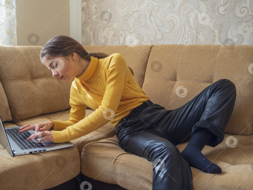 Скачать Красивая девушка-подросток в повседневной одежде сидит дома на диване с ноутбуком, реальные люди в реальной жизни фотосток Ozero