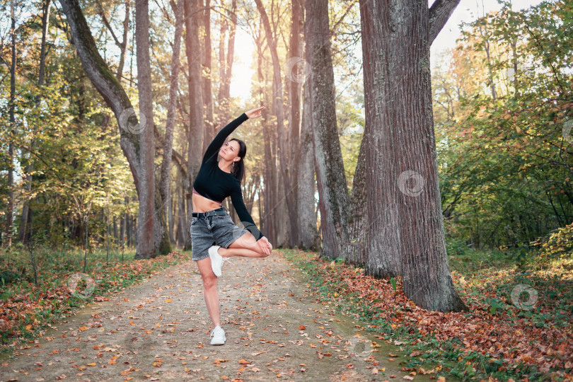Скачать Женщина, практикующая йогу, выполняет вариацию упражнения врикшасана, позу дерева, тренируясь в одиночестве в повседневной одежде солнечным осенним днем в парке, стоя на дорожке фотосток Ozero