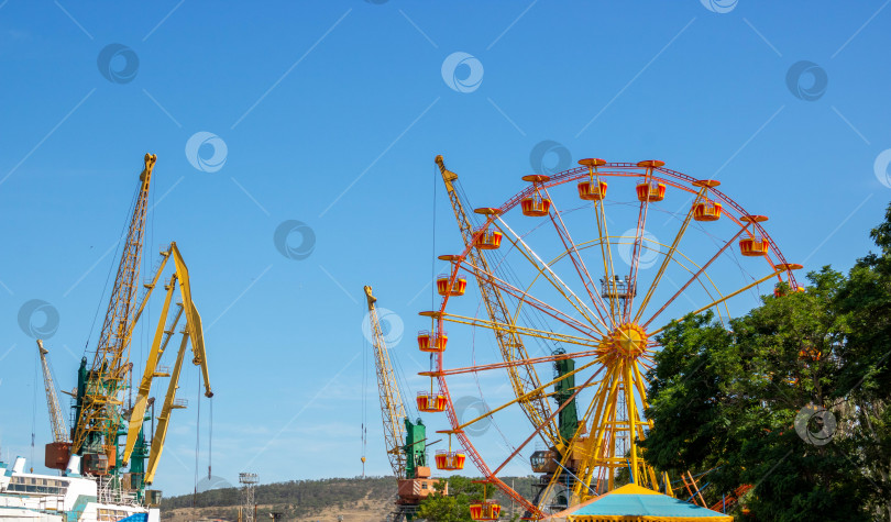 Скачать Летний день.Желтое колесо обозрения на фоне голубого неба, рядом с портальными кранами. фотосток Ozero