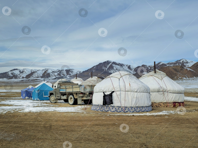 Скачать Монгольский юртовый лагерь в зимней степи. Рядом с юртами на фоне заснеженных гор стоит старый грузовик. фотосток Ozero