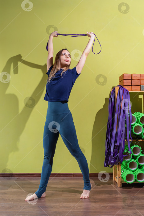 Скачать Молодая женщина тренируется в спортивной одежде с резинкой в руках, выполняя вытяжение поясничного и грудного отделов, поднимая руки вверх и отводя спину в плечевом суставе фотосток Ozero