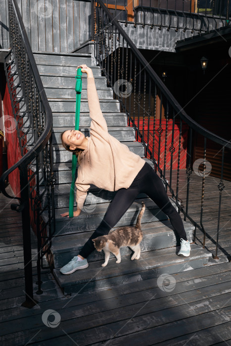 Скачать Довольная молодая женщина со своей кошкой занимается спортом на улице, выполняет упражнение Триконасана, позу треугольника с поясом для фитнеса, занимается йогой в спортивной одежде, стоя на порогах лестницы фотосток Ozero
