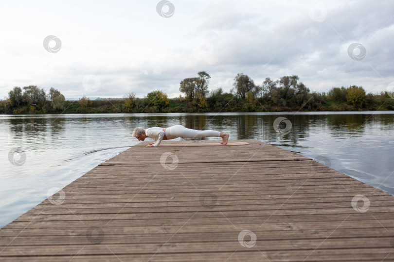 Скачать Зрелая женщина с короткой стрижкой в белом костюме занимается йогой на берегу реки. Чатуранга Дандасана, поза посоха с вытянутыми конечностями фотосток Ozero