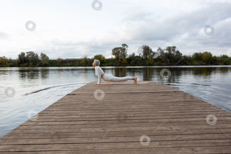 Скачать Зрелая женщина с короткой стрижкой в белом костюме занимается йогой на берегу реки. Бхуджангасана. Восходящая собака фотосток Ozero